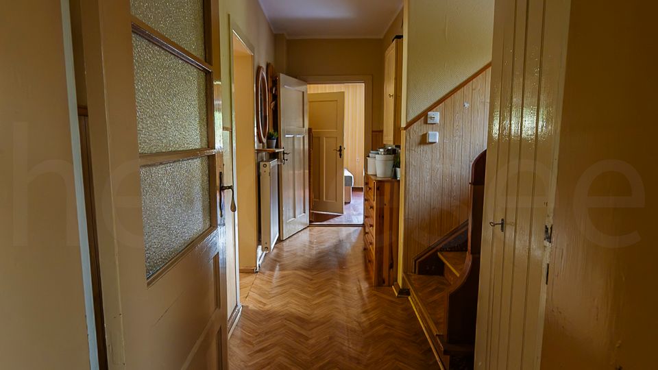 Schmaler Hausflur mit Fischgrät-Holzboden, cremefarbenen Wänden und einer Deckenleuchte. Links stehen Türen mit Milchglas, rechts ein Einbauschrank und der Beginn einer Holztreppe; am Ende führt eine offene Tür in einen weiteren Raum.
