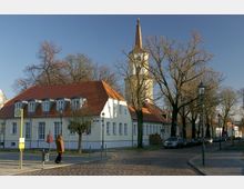Straßenszene in einer kleinen Ortschaft mit Kopfsteinpflaster, einem Gebäude mit rotem Ziegeldach und Dachgauben im Vordergrund sowie einer Kirche mit hohem Glockenturm im Hintergrund. Karge Bäume und Laternen säumen die Straße, neben der parkende Autos stehen.