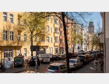 Straßenszene mit mehrstöckigen Wohnhäusern in Gelb- und Orangetönen, einer gepflasterten Straße und parkenden Autos am Straßenrand. Im Hintergrund ist ein Turm mit grünem Dach zu sehen, während Bäume mit frischem Laub den Straßenverlauf säumen. Ein Straßenschild zeigt "Boddinstraße" und "Isarstraße".