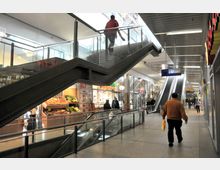 Einkaufsbereich einer U-Bahn-Station mit Geschäften auf beiden Seiten, einer Rolltreppe, einer Treppe und Hinweisschildern für den U-Bahn-Zugang. Im Vordergrund befinden sich ein Obst- und Gemüsestand sowie Personen, die durch den Bereich gehen.