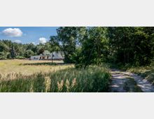 Ein ländlicher Weg führt durch eine Wiese mit hohen Gräsern und Bäumen im Hintergrund. Links steht ein modernes, einstöckiges Gebäude mit flachem Dach vor einem dichten Wald.