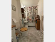 Kleines Badezimmer mit einer Badewanne, die durch einen Duschvorhang mit Blumenmuster verdeckt ist. Zu sehen sind eine Toilette mit Holzsitz, ein Waschbecken, ein schmaler Teppich auf weißen Fliesen, ein Holzbadezimmerschrank und ein Heizkörper an der Wand.