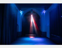 Ein dunkler Innenraum mit einem hohen, bogenförmigen Durchgang, beleuchtet von einem einzelnen, senkrechten Strahl aus farbigem Licht (rot und weiß) in der Mitte. Die Umgebung ist in blauem Licht getaucht, mit seitlich aufgehängten Vorhängen und einer leeren Sitzbank im Hintergrund.