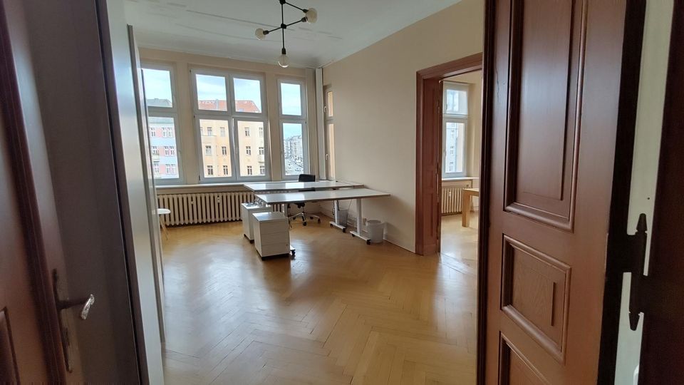 Heller Büroraum mit Fischgrätparkett, hohen Decken und großen Fensterfronten mit Blick auf gegenüberliegende Wohnhäuser. In der Mitte stehen zusammengestellte Schreibtische; dunkle Holztüren und ein Durchgang führen in einen weiteren Raum mit ähnlichen Fenstern und Heizkörpern.