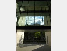 Blick durch eine moderne Passage mit Glasfassade und hellem Stein, die durch eine Überdachung unterbrochen wird. Im Hintergrund sind Bäume, eine Straße und ein Gebäude aus Stein zu sehen.