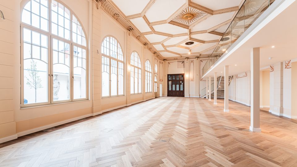 Heller, hoher Saal mit großen, bogenförmigen Sprossenfenstern entlang einer Wand und einem Fischgrät-Parkettboden. Die Decke ist reich mit Stuck und geometrischen Kassetten verziert; rechts befindet sich eine moderne Galerieebene mit Glasgeländer und Treppe.