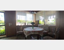 Gemütlicher Essbereich in einem holzverkleideten Raum mit großer Eckbank, gepolsterten Stühlen und einem ovalen Tisch mit bestickter Tischdecke. Die Fenster mit weißen Gardinen bieten Blick auf einen grünen Garten.