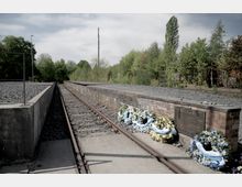Gleisanlage mit einem Mahnmal aus Backstein, darauf eine Gedenktafel und mehrere Gedenkkränze mit blauen und weißen Schleifen. Im Hintergrund sind Bäume und Büsche zu sehen, die Umgebung wirkt ruhig und grün.
