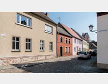 Eine schmale, gepflasterte Straße mit einfachen Wohngebäuden in verschiedenen Farben, darunter verputzte und Ziegelwände. Im Hintergrund ist der Turm einer Kirche sichtbar, und einige geparkte Autos stehen entlang der Straße. Straßenschilder zeigen die "Pfarrgasse" und die "Vogelsangstraße".