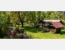 Ein kleiner Wasserkanal in einer grünen Parklandschaft, auf dem eine Familie in einem roten Kanu fährt. Rechts befinden sich eine überdachte Holzstruktur und ein Holzfass mit Dach, umgeben von Bäumen und Rasenflächen.