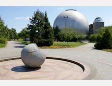 Ein moderner Park mit gepflasterten Wegen, einem großen kugelförmigen Gebäude im Hintergrund (vermutlich ein Planetarium) und einer kleineren, zylindrischen Struktur daneben. Im Vordergrund befindet sich eine Steinskulptur auf einem kreisförmigen Steinpodest, umgeben von Bäumen, Rasenflächen und Blumenbeeten.