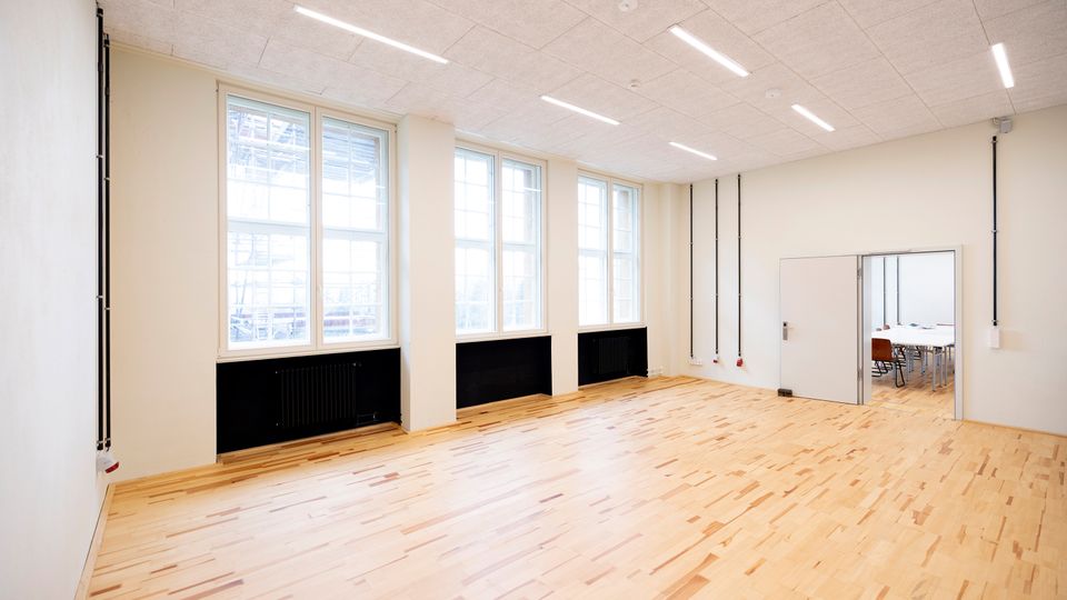 Heller, leerer Innenraum mit hellem Holzfußboden, hohen Fenstern entlang einer Wand und schwarzen Heizkörpern darunter. Abgehängte Decke mit langen LED-Lichtleisten sowie eine Türöffnung zu einem angrenzenden Raum mit Tischen und Stühlen.