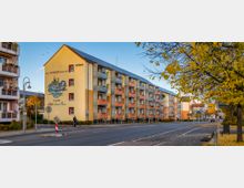 Wohngebäude an einer Straße in einem städtischen Bereich mit moderner Fassade in Gelb- und Orangetönen, auf der ein Wandgemälde sowie die Aufschrift „1265 SCHWEDT/ODER 2015“ sichtbar sind. Im Vordergrund befinden sich Gehwege mit herbstlich gefärbten Bäumen und Straßenschilder.