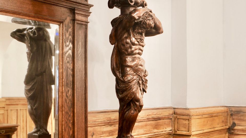 Detailansicht eines reich verzierten Holzinterieurs mit großem Rahmen, Spiegel und geschnitzter Figur als Stütze. Im Hintergrund sind helle Wände und holzvertäfelte Wandpaneele zu sehen.