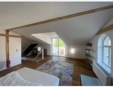 Ein Dachgeschossraum mit schrägen Decken, Holzbalken und Holzfußboden. Der Raum umfasst ein Bett im Vordergrund, ein großes Fenster mit Blick nach draußen, ein kleines rundes Fenster und ein Glastürregal an der rechten Wand.