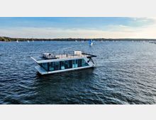 Ein modernes Hausboot mit großflächigen Fenstern und einer Dachterrasse mit Sitzmöbeln treibt auf einem See, umgeben von zahlreichen Segelbooten und einer bewaldeten Uferlandschaft im Hintergrund.