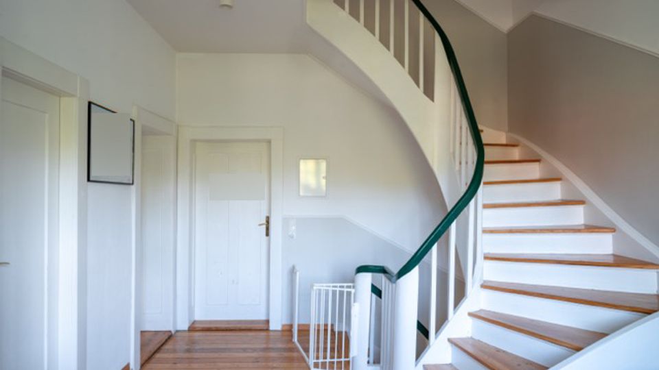 Heller Flur in einem Wohnhaus mit Holzdielenboden und weißen Türen an der linken Wand. Rechts führt eine geschwungene Treppe mit Holzstufen, weißem Geländer und dunkelgrünem Handlauf nach oben.