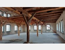 Offener Dachbodenraum in einem historischen Gebäude mit unverputzten Backsteinwänden, sichtbaren Holzbalken, Stützpfosten aus Holz und einer Reihe von großen, bogenförmigen Fenstern. Der Holzfußboden ist teilweise beschädigt, und das Licht fällt durch die Fenster in den leeren Raum.