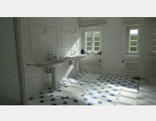 Helles Badezimmer mit weißen Fliesenwänden und blau-weißen Bodenfliesen. Zwei Waschbecken mit Spiegeln, ein Fenster mit Jalousien und ein Heizkörper sind ebenfalls zu sehen.