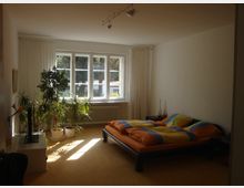 Ein Schlafzimmer mit einem großen Fenster, durch das Tageslicht einfällt. Der Raum ist hell eingerichtet, mit einem Doppelbett und mehreren Topfpflanzen neben einem Heizkörper unter dem Fenster.