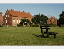 Eine grüne Wiese mit einer einfachen Holzbank im Vordergrund, im Hintergrund zwei Backsteinhäuser mit roten Dächern, umgeben von Bäumen und Sträuchern, bei klarem Himmel.