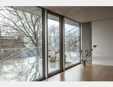 Ein minimalistischer Innenraum mit heller Holzbodenfläche und raumhohen Fenstern, die auf eine Außenterrasse mit Metallgeländern und Außentreppen blicken. Draußen sind kahle Bäume, eine ziegelverkleidete Wand und eine regennasse Oberfläche zu sehen, innen steht eine Vase mit trockenen Zweigen.