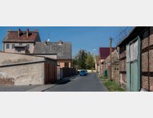 Straße in einem Wohngebiet mit alten Gebäuden, teils aus Ziegelmauerwerk und Putz, sowie Garagen mit roten Toren auf der linken Seite. Im Hintergrund sind Wohnhäuser mit Schrägdächern und ein geparkter blauer PKW sichtbar, umgeben von Bäumen und einem klaren blauen Himmel.