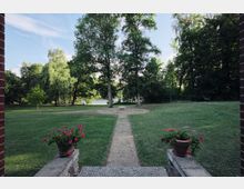 Blick von einem gepflasterten Eingangsbereich mit Blumenkübeln auf eine gepflegte Rasenfläche mit einem Weg, der zu einem runden Sandplatz mit einer Bank führt; im Hintergrund stehen große Bäume vor einem See.