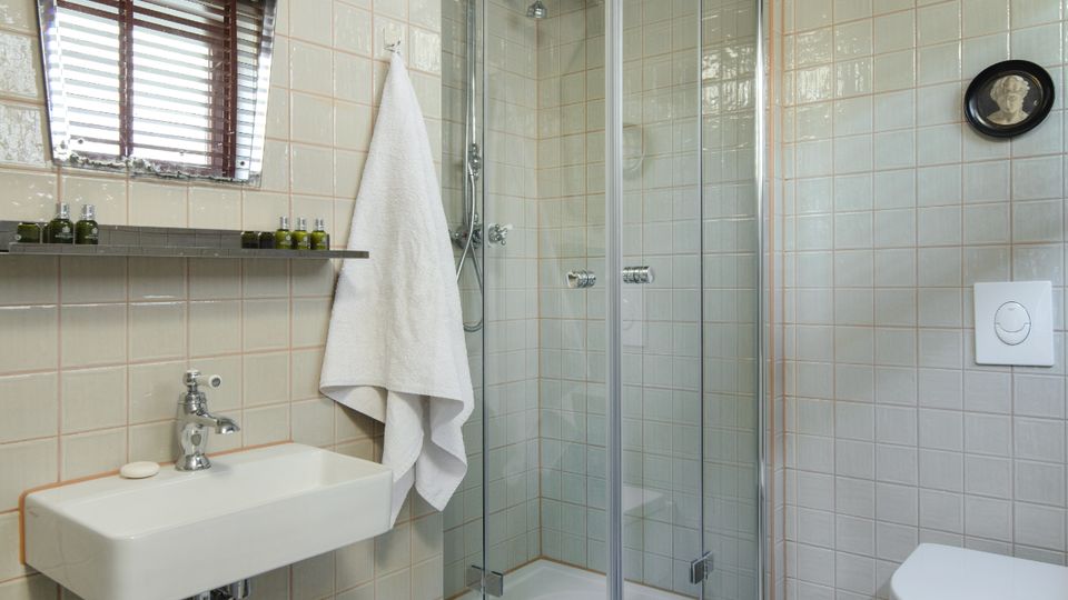 Modern bathroom with white tiled walls, a concrete ceiling, and a glass-enclosed corner shower. A wall-mounted sink beneath an angular mirror, a hanging towel, and a wall-hung toilet sit on a smooth light-gray floor.