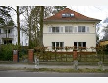 Zweistöckiges Wohnhaus mit gelber Fassade und einem roten Ziegeldach, umgeben von kahlen Bäumen im Vorgarten und einer Hecke hinter einem Metallzaun mit Backsteinpfosten. Im Vordergrund befindet sich ein Bürgersteig entlang einer Straße.