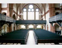 Innenraum einer Kirche mit Backsteinbögen, hohen Decken und großen Fenstern mit Spitzbögen und Maßwerk. Dunkle Holzempore und Sitzbänke dominieren den Raum, der zentral durch einen Mittelgang geteilt ist.