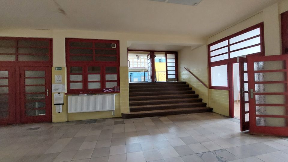 Eingangsbereich eines Gebäudes mit cremefarbenen Wänden, roten Rahmen aus Holz und Glaselementen an Türen und Fenstern. Eine breite Treppe mit dunklem Holz führt zu einer weiteren Glastür, die ins Freie zeigt.