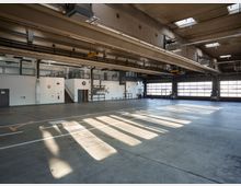 Große, helle Industriehalle mit Betonboden, hohen Decken und Oberlichtern. An den Seiten befinden sich weiße Wände, eine Treppe zu einem Zwischenstockwerk mit Geländer sowie große Rolltore mit Fenstern, die Tageslicht hereinlassen.