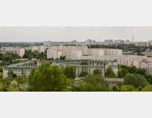 Stadtansicht mit mehreren Wohnblocks, darunter grüne und beige Plattenbauten, umgeben von Bäumen und Grünflächen. Im Hintergrund sind weitere Hochhäuser und eine weitläufige urbane Landschaft zu sehen.