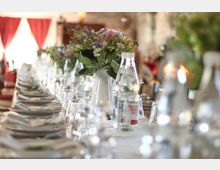 Langer, festlich gedeckter Tisch mit weißen Tischdecken, Geschirr, Gläsern und Wasserflaschen. In der Mitte des Tisches stehen Blumensträuße in weißen Vasen; im Hintergrund rote Vorhänge und ein rustikales Ambiente.