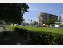 Eine städtische Szene mit einer Rasenfläche im Vordergrund, flankiert von Hecken und Sitzbänken, und einem mehrstöckigen Bürogebäude mit der Aufschrift "Eternit" im Hintergrund. Mehrere Flaggen stehen vor der Straße, auf der Autos und Lieferwagen fahren.