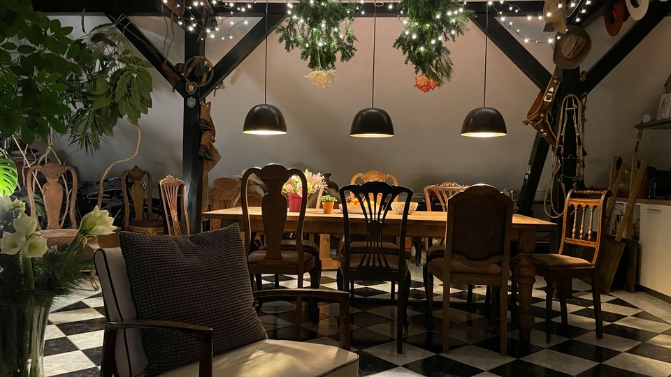 Gemütlicher Innenraum mit sichtbaren schwarzen Dachbalken, Lichterketten und hängendem Grün, darunter ein langer Holztisch mit unterschiedlichen Stühlen. Schwarz-weiß karierter Fliesenboden, mehrere Pendelleuchten sowie Pflanzen und dekorative Gegenstände an Wänden und Regalen.