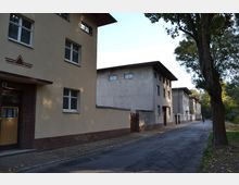 Straßenszene mit mehreren zweigeschossigen Wohngebäuden im Stil der Nachkriegszeit, teils verputzt und teils mit Backsteinsockeln. Die Straße ist schmal, gesäumt von Bäumen und leicht mit Laub bedeckt; im Hintergrund sind Menschen und Fahrräder erkennbar.