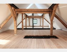 Dachbodenraum mit sichtbaren Holzbalken und hellem Holzfußboden, ergänzt durch ein kleines Fenster in der Mitte an der Stirnwand. Die weißen Wände und schrägen Decken betonen die offene und minimalistische Gestaltung.