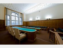 Raum mit klassischer Holzvertäfelung und hohen Fenstern mit Rundbogen im oberen Bereich. Es steht ein großer Konferenztisch mit beige gepolsterten Stühlen in der Mitte, flankiert von weiteren Stühlen an den Wänden; die Decke ist mit geometrischen Mustern verziert.