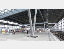 Weitläufiger und moderner Bahnhof mit überdachtem Bahnsteig, großen Glasfassaden, massiven Stahlträgern und Beschilderung, die "Berlin Südkreuz" ausweist. Der Bahnhof enthält Sitzbereiche, Geschäfte und ein Fahrrad, das von einer Person geschoben wird.