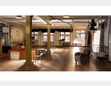 Großer, offener Raum mit hohen Decken, durchsetzt mit gelben Metallsäulen und ausgestattet mit Holzboden. Der Raum enthält diverse Sitzmöbel, Arbeitstische, Fotografie- oder Filmgeräte sowie große Fenster, durch die Tageslicht einfällt.