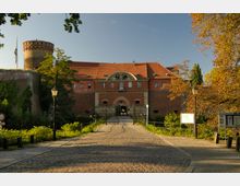 Historisches Backsteingebäude mit rotem Ziegeldach und Rundturm, umgeben von Bäumen und einem gepflasterten Weg, der zu einem Torbogen mit schmiedeeisernem Gitter führt. Im Vordergrund Laternen und Hinweisschilder entlang des Weges.