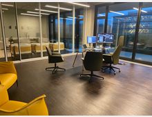 Modernes Büro mit Glaswänden und Holzboden, ausgestattet mit Schreibtischen, Bürostühlen und zwei Monitoren. Im Hintergrund ist eine Lounge mit gelben und weißen Sofas sowie ein Ausblick auf ein benachbartes Bürogebäude durch große Fenster zu sehen.