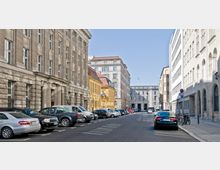 Straßenszene mit historischen und modernen Gebäuden: Links ein großes Steingebäude mit Säulenfassade, daneben ein gelbes Haus mit roten Dachziegeln. Rechts moderne und klassische Bauten. Entlang der Straße sind Autos geparkt.