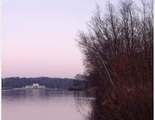 Blick auf einen See bei Sonnenuntergang mit kahlen Bäumen am rechten Ufer. Im Hintergrund ist ein weißes, historisches Gebäude am gegenüberliegenden Seeufer zu sehen, umgeben von Wald.