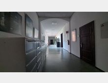 Langer, schmaler Flur in einem Büro- oder Verwaltungsgebäude mit weißen Wänden, einer abgehängten Decke und einem glatten Boden. Auf der linken Seite befinden sich Metall-Aktenfächer, während rechts Türen und Wandanzeigen sichtbar sind; der Flur ist spärlich beleuchtet und endet in einem dunkleren Bereich.