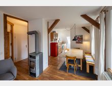 Wohnbereich mit offenem Übergang zur Küche, ausgestattet mit Holzfußboden, einem Kaminofen, einem Esstisch aus hellem Holz und rustikalen Holzbalken an der Decke. Im Hintergrund ist eine Küche mit Holzschränken, einem roten Kühlschrank und einem Dachfenster zu sehen.