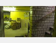 Ein einfacher Kellerraum mit gelb gestrichenen Wänden und unverputzten Ziegelabschnitten. Im Raum befinden sich gestapelte Pappkartons, Säcke und ein Besen, sowie eine Treppe mit Holzwangengeländer, die nach oben führt.