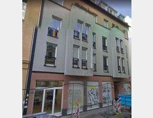 Mehrstöckiges Wohn- und Geschäftsgebäude mit grauer und rosafarbener Fassade. Die unteren Schaufensterbereiche sind mit Graffiti besprüht, darüber befinden sich kleinere schwarze Balkone vor den Fenstern. Vor dem Gebäude ist eine abgesperrte Baustelle sichtbar.